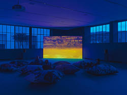 Video installation at night in a gallery space surrounded by large pillows.