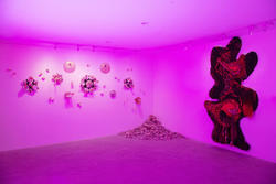 Image of a gallery in pink fluorescent light. Hung sculptures of fibers and beads on the wall and a pile of sand in the corner.