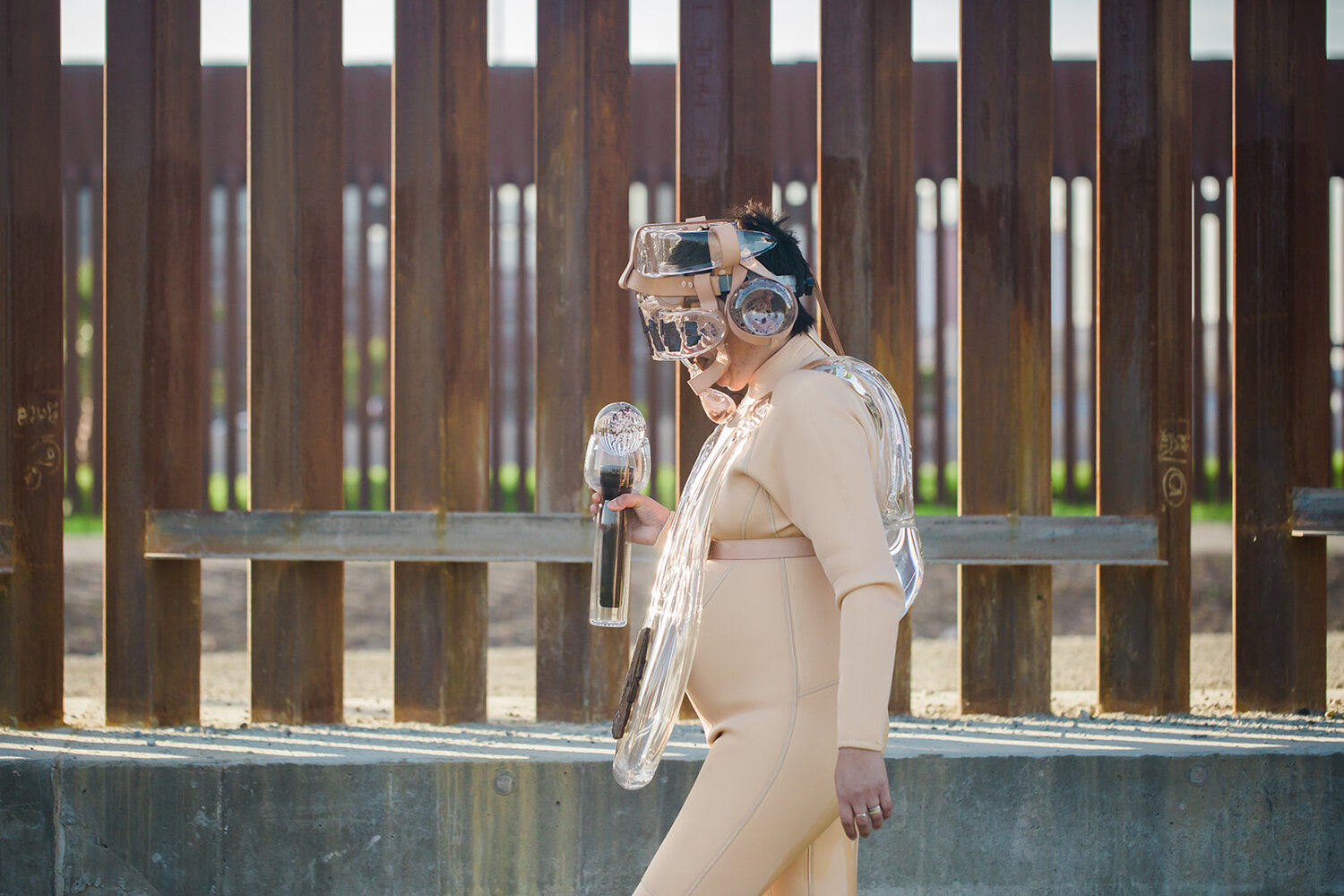 Alum Tanya Aguiñiga in a suit and mask made of flesh-toned neoprene, glass and metal, walking the length of the border wall in the southern US.