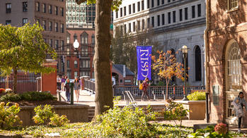 a blue flag with the letters RISD hangs in downtown Providence as students walk near green and gold foliage