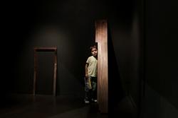 Art piece by Rosa Park, MFA 2016. Photograph of an unamused boy standing next to dimly lit wooden art display.