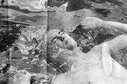 black and white image of a person's face, part of their torso, and arms extended above their head, floating on water