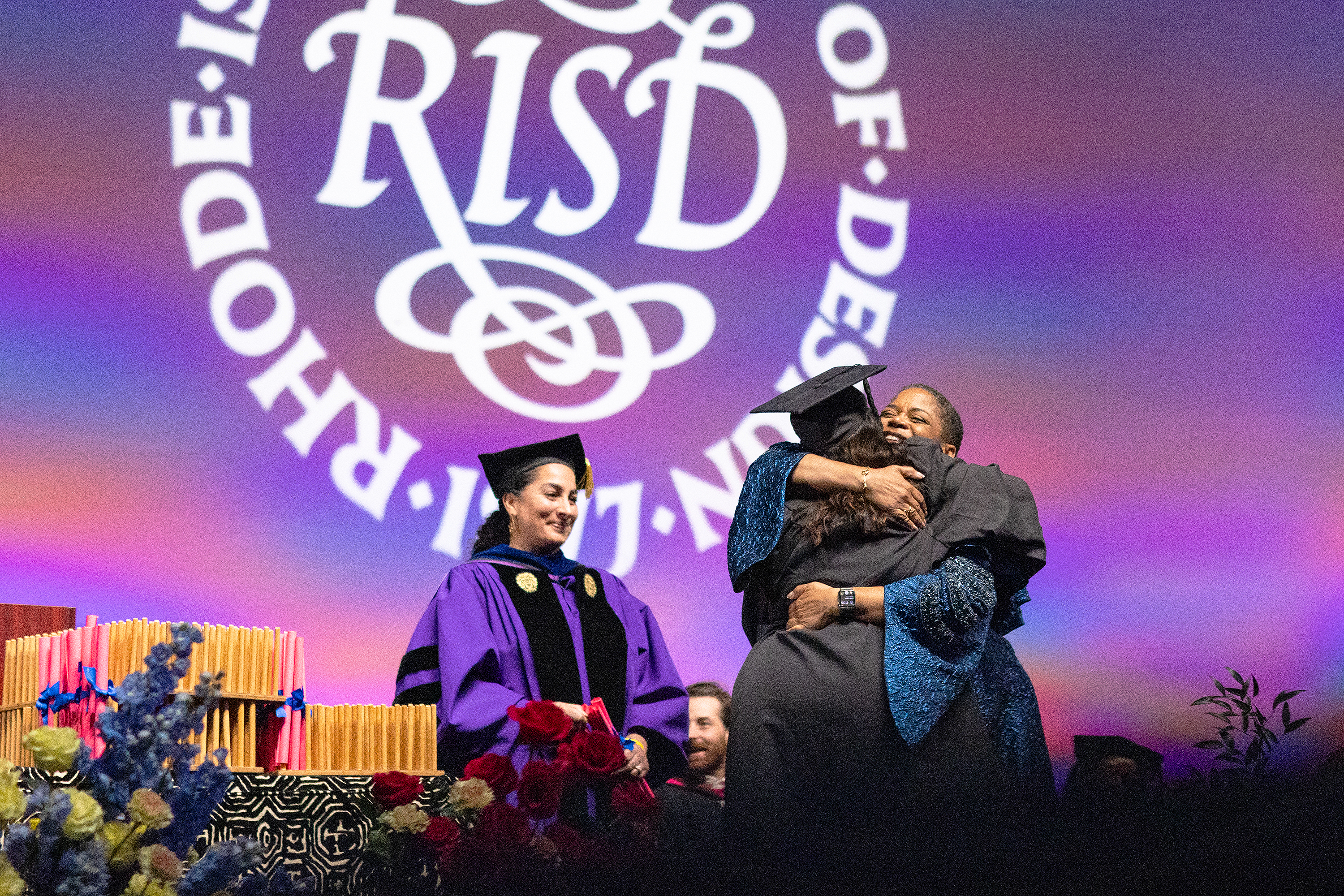 Provost Ghadessi looks on as President Williams hugs a graduate student at Commencement 2024