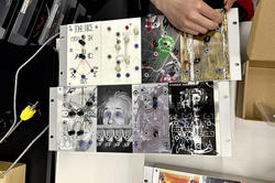 a hand manipulates a device featuring eight punk zine-style images, including one of Albert Einstein, with holes for audio cables prominent along the surface