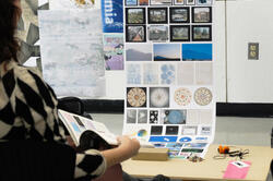 a student's hand reaches towards a long sheet with many small, square images hangs from a wall, surrounded by other printed works