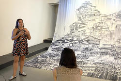 A person in a floral dress speaking near a large print drawing of many buildings