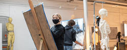 a student, facing the camera, draws on a large piece of newsprint next to a skeletal model and another drawing student