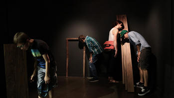 people in a dark room standing in a listening posture next to wooden structures