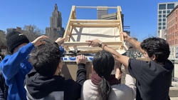 four students lift up a wooden structure, with the downtown Providence skyline in the distance