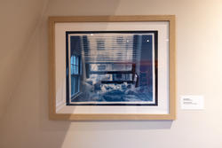 a framed photograph of a window inside a dark, blue-lit room, covered by open horizontal blinds and looking out onto the city