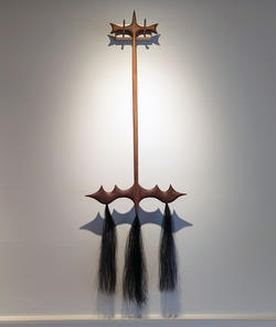 a broom-like sculpture with three tassels dangling from the lower end, hung from a gallery wall